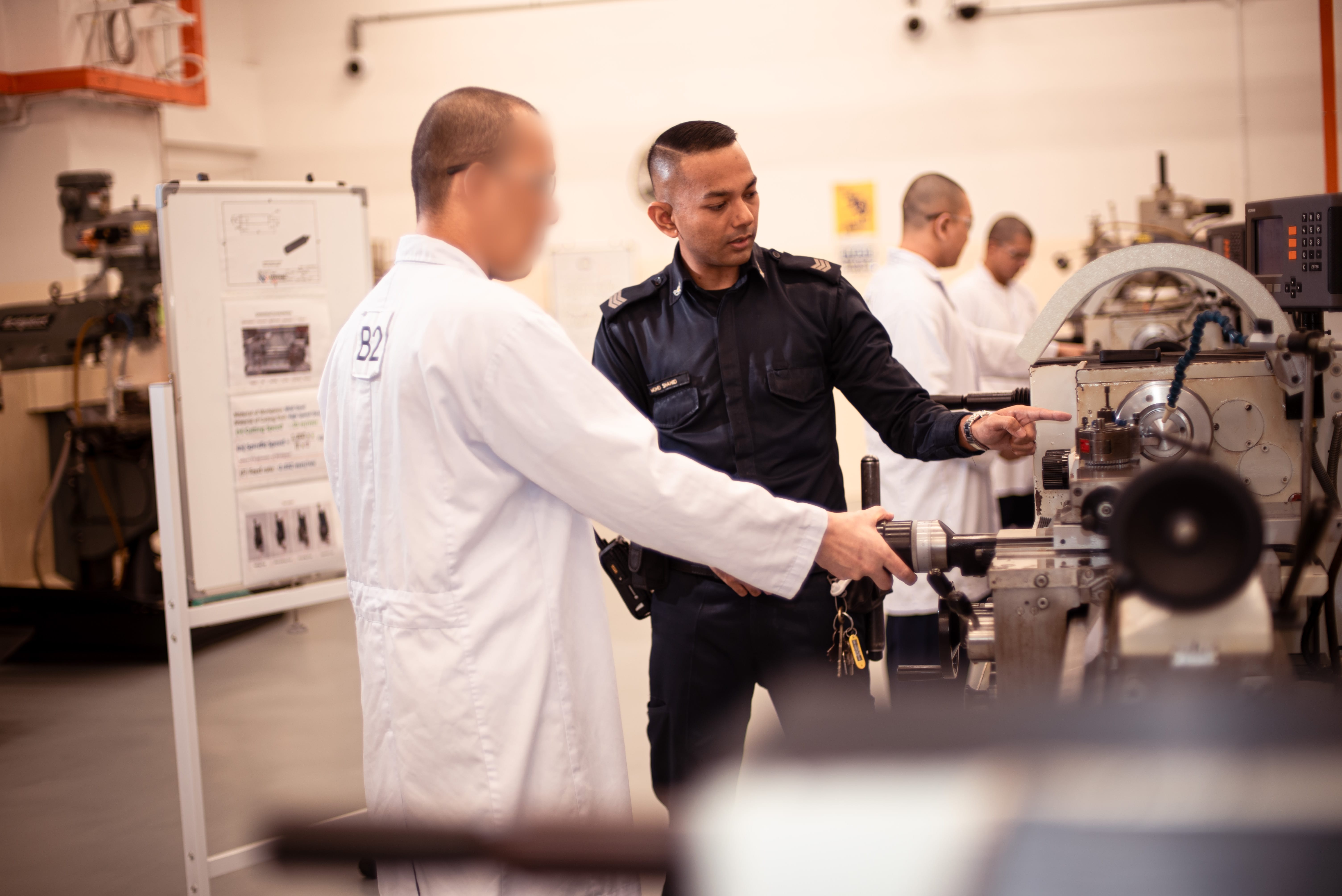 A correctional officer in a dark blue uniform provides hands-on guidance to an inmate in a white workshop uniform while operating a large metalworking machine in a vocational training workshop. The officer points to a specific part of the machine as the inmate follows the instruction with one hand on the equipment's controls. In the background, several other inmates wearing similar white uniforms are engaged in practical training at additional machines, creating the atmosphere of an active industrial classroom. A whiteboard displaying instructional materials stands nearby, reinforcing the structured learning environment. The image conveys vocational skills training, mentorship, and rehabilitation through practical workplace education within a correctional setting.